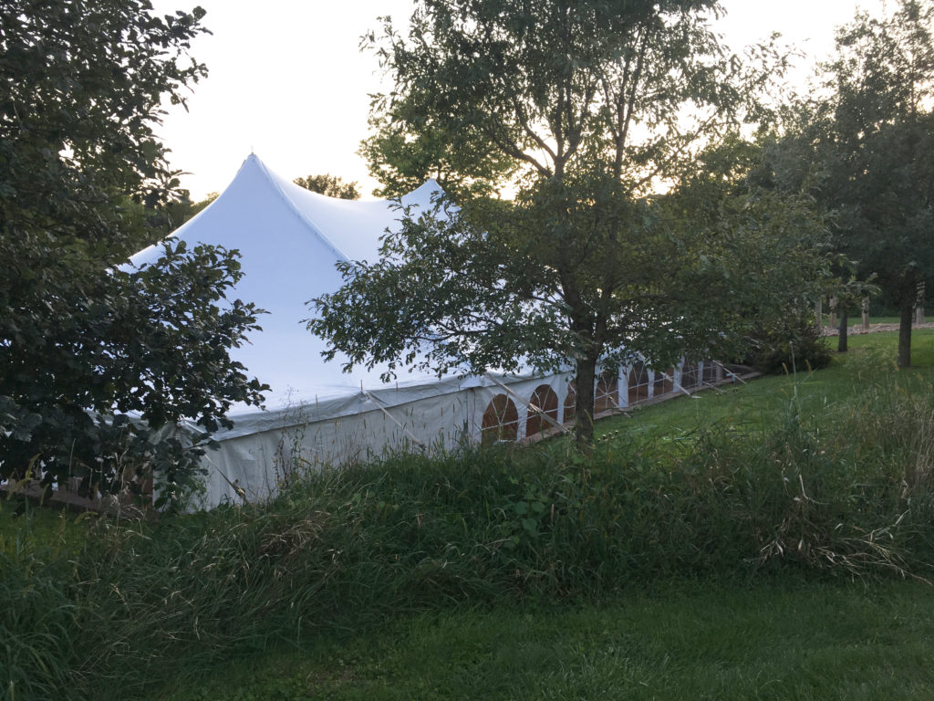 View through trees of the 40' x 60' white rope and pole wedding tent at Harvest Preserve