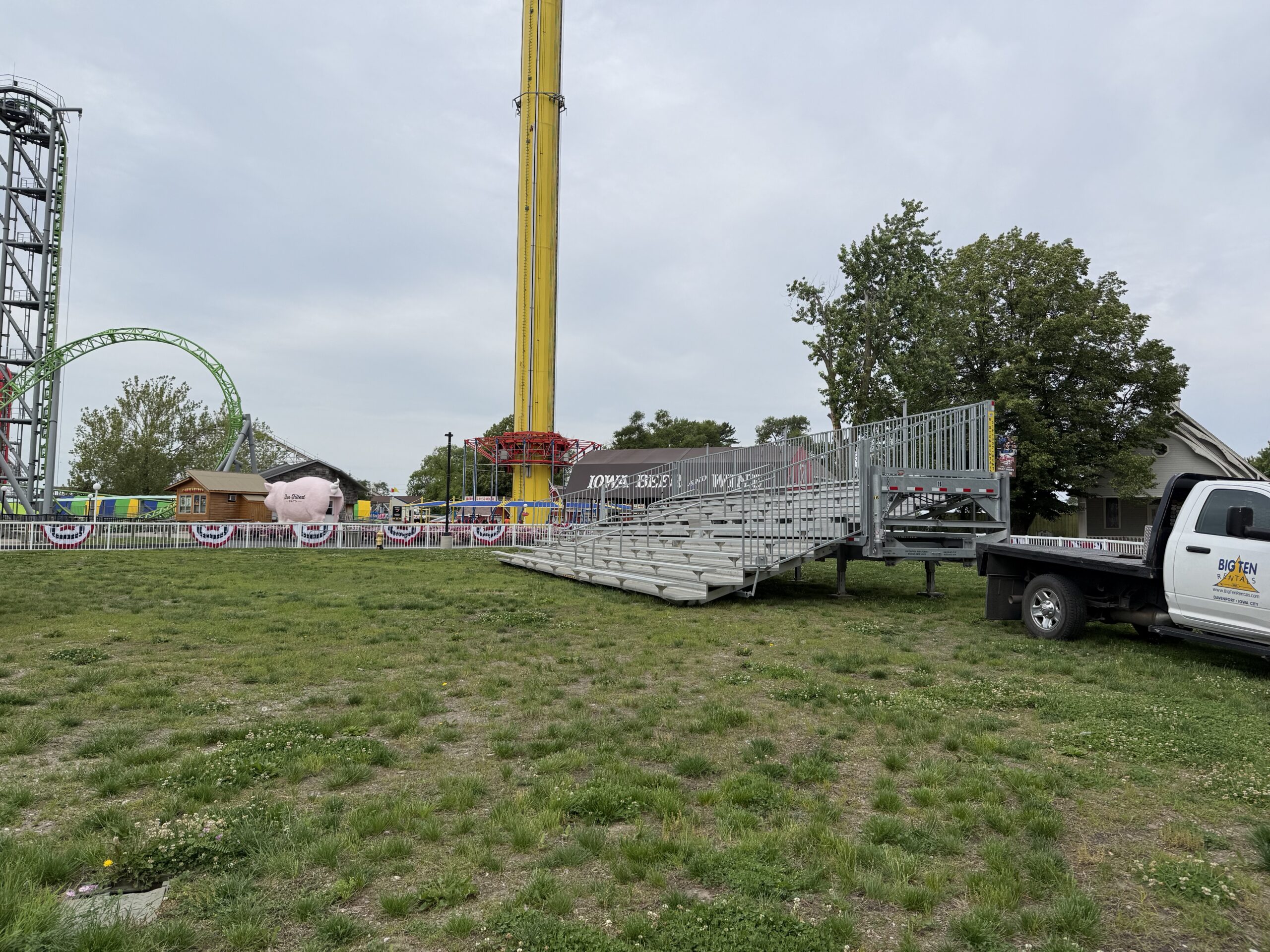 Angled view with Big Ten Rentals truck, Iowa Beer & Wine building, yellow drop tower, green roller coaster, and pink pig landmark in the background at Adventureland Park in Altoona, Iowa.