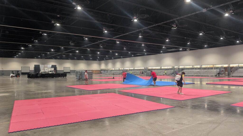 Crew laying out colorful interlocking foam mats on the competition floor with spectator bleachers visible in the background.