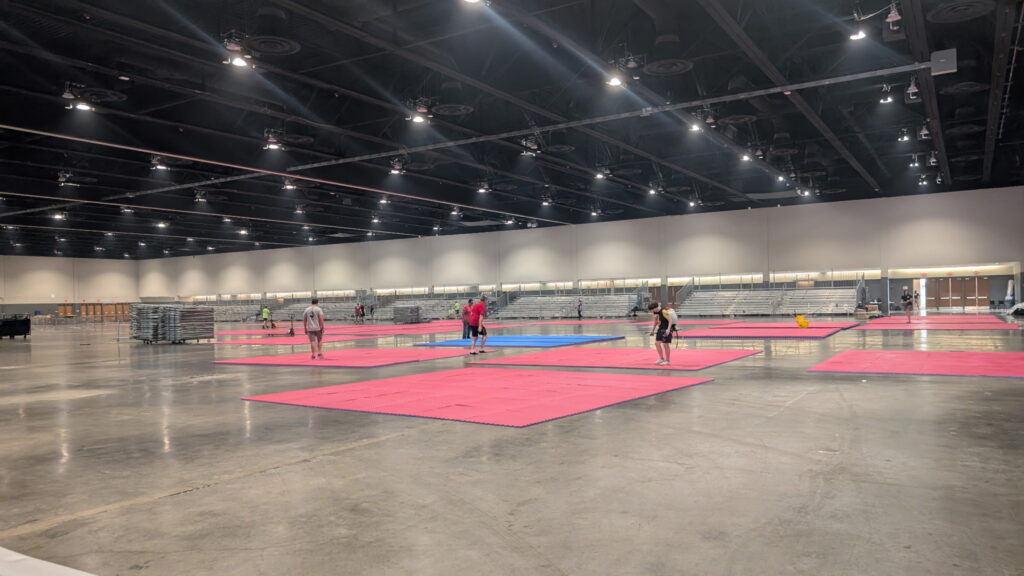 Wide view of the Schaumburg Convention Center hall with portable bleachers installed and pink/blue foam mats being laid for WTSDA martial arts competitions.